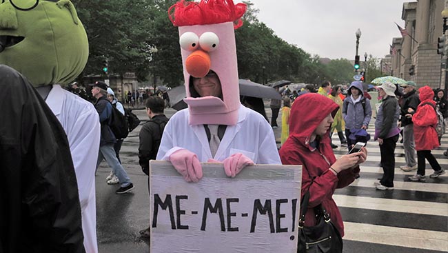 Protestor dressed in costume as muppet charcter "Beaker" holding a sign that reads: "ME-ME-ME!"